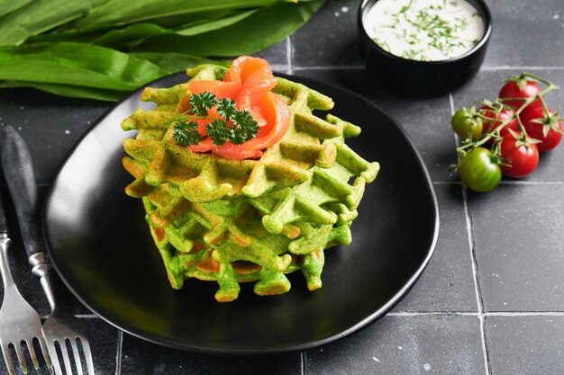 Green Belgian waffles Spinach or wild garlic or pesto waffles with red salmon and cream sauce on old black tile table background Delicious breakfast snack brunch on dark background