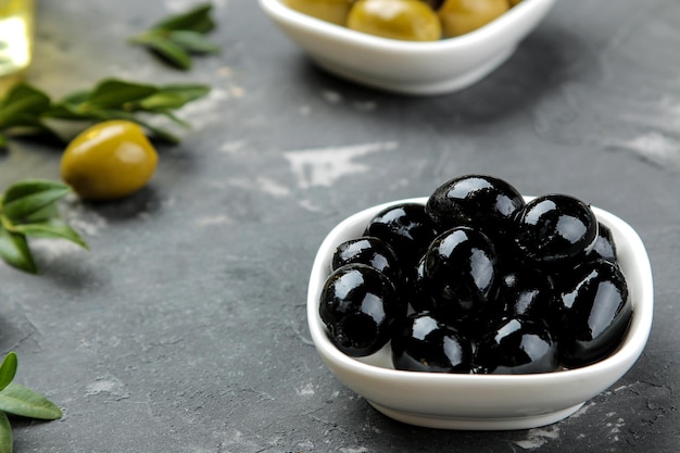 Green and black olives in a white ceramic bowl with leaves on a dark graphite background. close-up