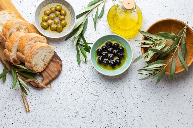 Green and black olives with olive oil in a glass bottle, olive tree sprigs and cut fresh ciabatta bread on wooden cutting board. White rustic background, healthy mediterranean food, space for text