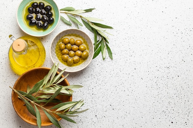 Green and black olives with olive oil in a glass bottle, olive tree sprigs and cut fresh ciabatta bread on wooden cutting board. White rustic background, healthy mediterranean food, space for text