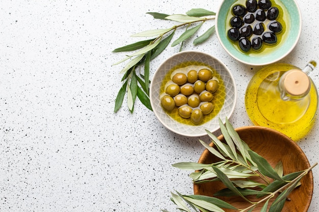 Green and black olives with olive oil in a glass bottle, olive tree sprigs and cut fresh ciabatta bread on wooden cutting board. White rustic background, healthy mediterranean food, space for text