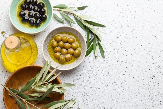 Green and black olives with olive oil in a glass bottle, olive tree sprigs and cut fresh ciabatta bread on wooden cutting board. White rustic background, healthy mediterranean food, space for text