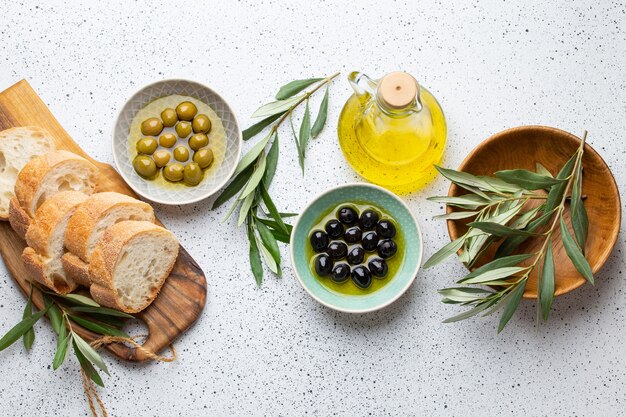 Green and black olives with olive oil in a glass bottle, olive tree sprigs and cut fresh ciabatta bread on wooden cutting board. White rustic background, mediterranean food concept, top view