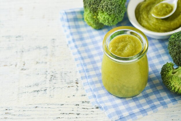 Green broccoli baby food in white bowl and jar on table Green baby food Child first feeding concept Baby Natural Food Production and menu of baby food Selective focus