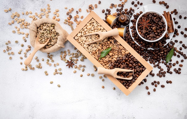 Green and brown decaf unroasted and dark roasted coffee beans in wooden box with scoops setup on white concrete.