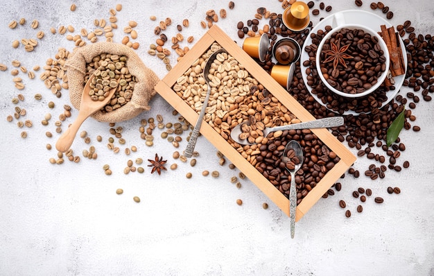 Green and brown decaf unroasted and dark roasted coffee beans in wooden box with scoops setup on white concrete.