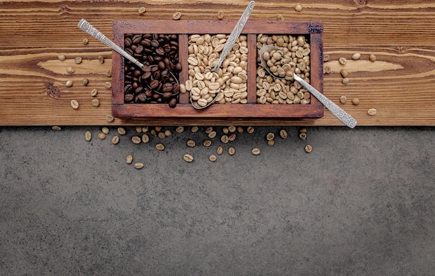 Green and brown unroasted and dark roasted coffee beans in wooden box with spoons setup on dark concrete background