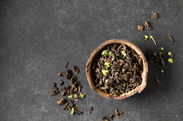 Green dry tea leaf with sausep fruit in a coconut bowl with scattered leaves, gray background, close-up, top view