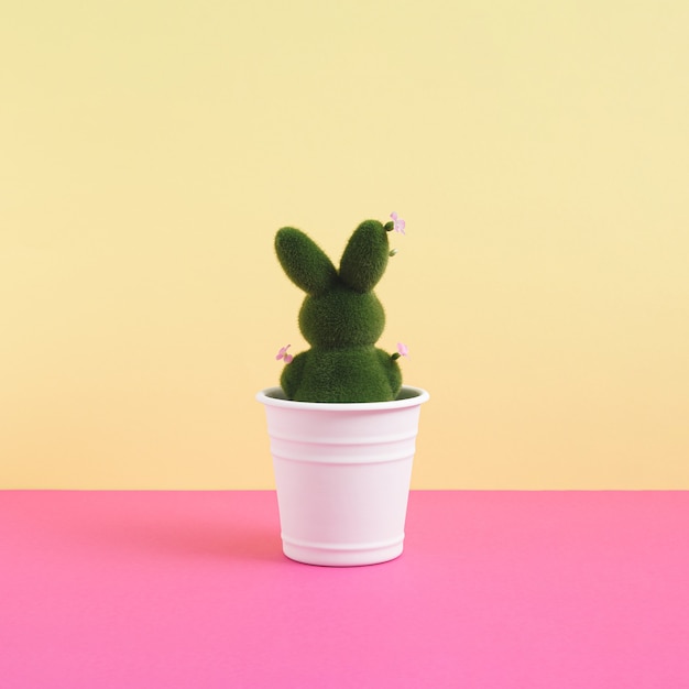 Green easter bunny decorated with flowers in flowerpot on vibrant yellow and pink background. Minimalist concept. Square with copy space.