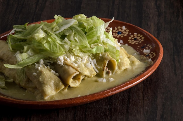 Green Enchiladas with lettuce served in a clay dish on a wooden table.