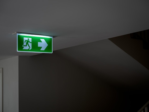 Green fire escape sign hang on the ceiling in the dark building near the stairway Emergency fire exit sign warning plate with running man icon and arrow to the right way with copy space