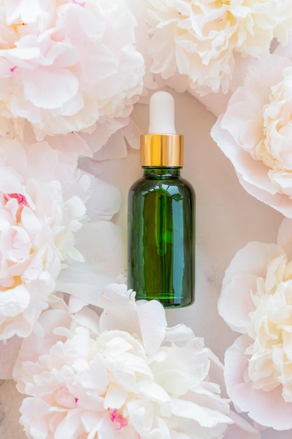 A green glass bottle with a serum essential oil or other cosmetic product against a background of pink peonies Natural organic cosmetic packaging skin care concept