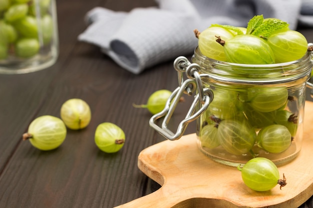 Green gooseberries in glass jar on cutting board. Gray napkin and gooseberries on table. Dark wooden background. Top view