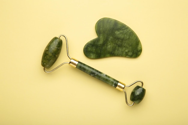 Green jade roller and gua sha stone for facial massage and on beige background Home beauty and selfcare accessories Face roller for anti age wrinkle treatment Top view flat lay