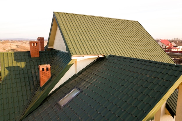 Green metal shingled house roof with attic plastic window and brick chimney.