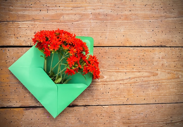 Green paper envelope with fresh garden red lychnis flowers on aged wooden boards background. Festive floral template. Greeting card design. Top view.