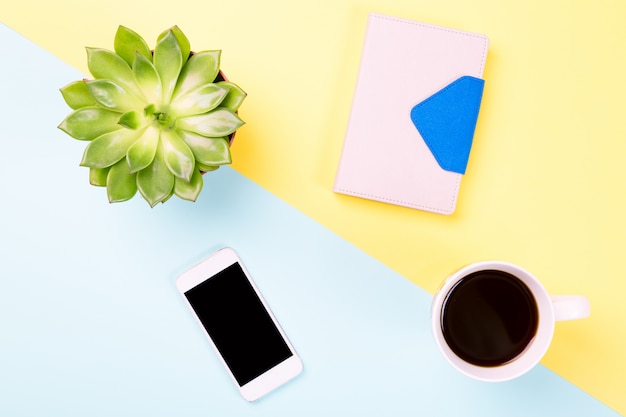 Green plant in a pot, cup of coffee, notebook and modern mobile phone on blue and yellow pastel surface.