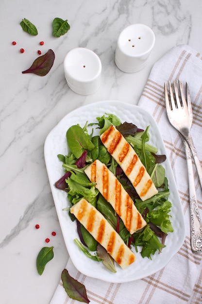 Green salad with fried halloumi cheese in a white plate on a marble table, top view