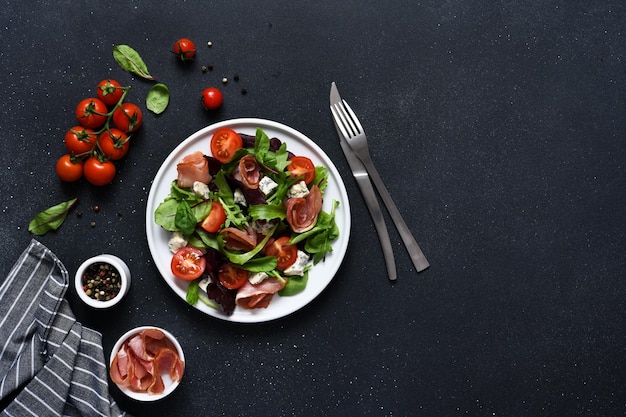 Green salad with prosciutto tomatoes and blue cheese on a black background top view