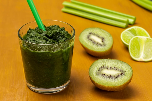 Green smoothie and straw in glass Kiwi and lime slices celery stalks on table