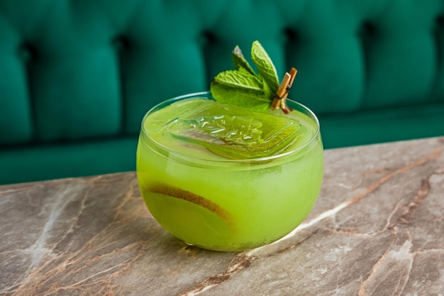 A green sour alcoholic fruit cocktail with kiwi served with a big ice cube in a lowball rocks glass garnished with mint