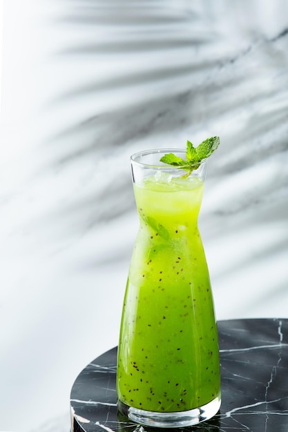 Green summer lemonade in jug on light background with shadow of branch tropic plant. Lemonade with kiwi and ice