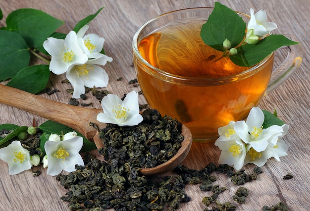 green tea with jasmine. dry green tea leaves with jasmine flowers in a wooden spoon and a cup of tea on a wooden table