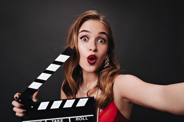 Photo greeneyed woman with red lipstick holding clapperboard shocked blonde lady in silk top looking into camera with surprise and posing on blacl background