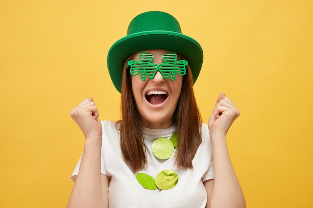 Greenthemed event Irish culture celebration Overjoyed Caucasian woman wearing green leprechaun hat and clover glasses standing isolated over yellow background with clenched fists