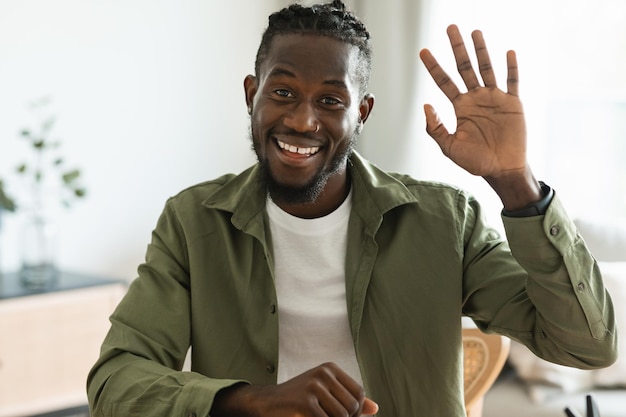 Greeting concept Friendly black man waving at camera and smiling saying hi while having video call at home