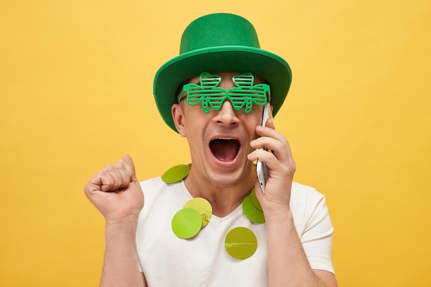 Greetings for Irish celebrations St Patricks Day mythology Amazed Caucasian man wearing green leprechaun hat and clover glasses standing isolated over yellow background talking on mobile phone