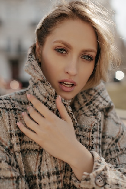 Grey-eyed blonde attractive woman in warm stylish checkered tweed coat looks into camera and walks outdoors