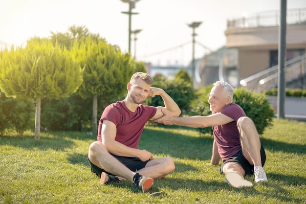 Grey-haired man touching bearded male biceps, both sitting on grass