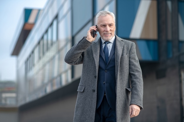 Grey-haired man wearing a grey coat talking on the phone standing near the office