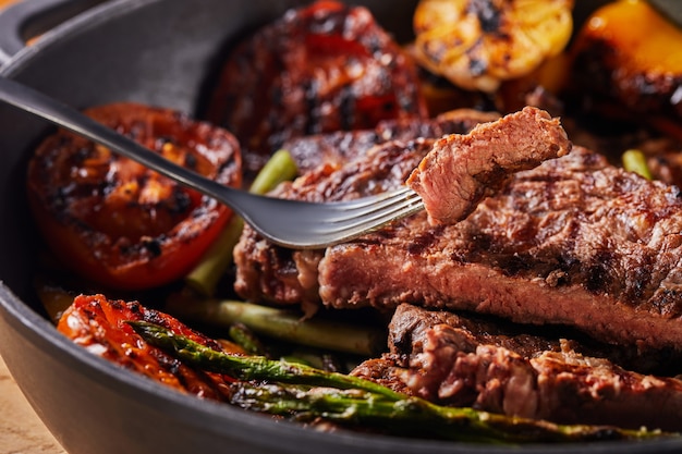 Grilled Beef steak in a black pan, a piece of chopped on a fork, with baked vegetables - tomatoes, asparagus, garlic and peppers