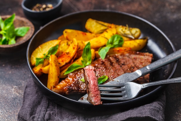 Grilled beef steak with potatoes and basil in a black plate on dark background.