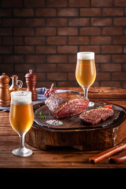 Grilled cap rump steak on wooden cutting board with two sweaty cold tulipa glasses of beer. Wooden table and bricks wall background (Brazilian picanha).