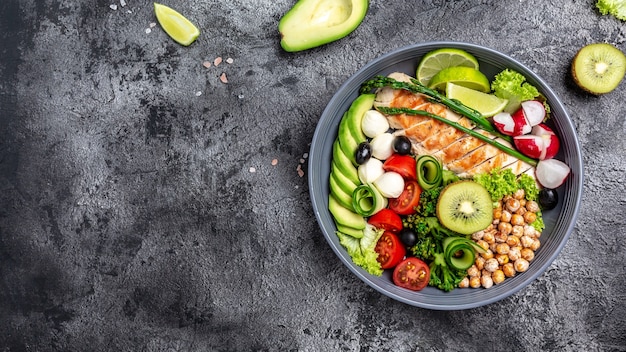 Grilled chicken, avocado, asparagus, chickpeas, broccoli, radish, chicken, cucumber, tomatoes, olives, mozzarella buddha bowl on dark background, top view. Delicious balanced food concept