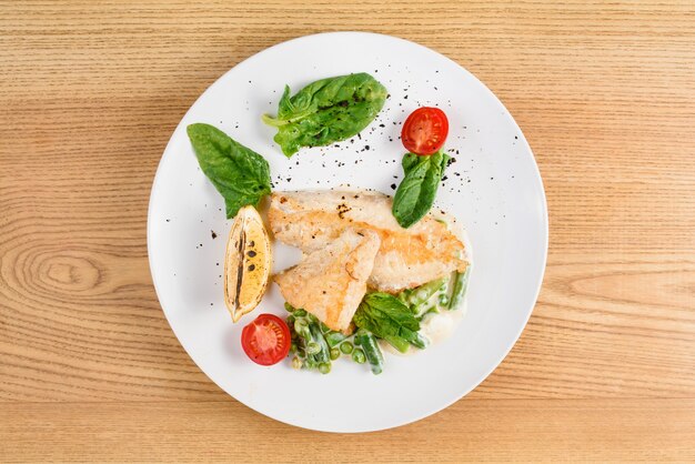 Grilled chicken fillet with basil, cherry tomatoes, lemon, peas, sauce on white round plate on wooden table