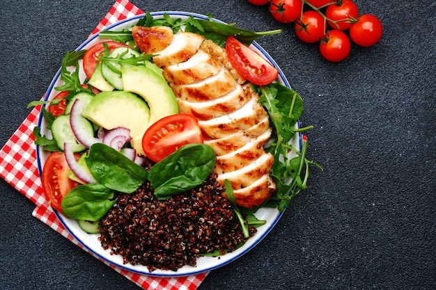 Grilled chicken meat red quinoa and fresh vegetable salad with tomato avocado arugula and spinach Healthy detox ketogenic diet food Black background top view