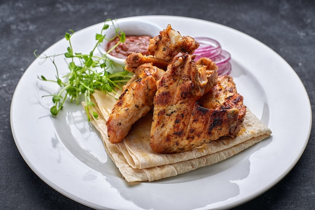 Grilled chicken wings with barbecue sauce, pita bread, microgreen and onion rings, on a white plate, against a dark table