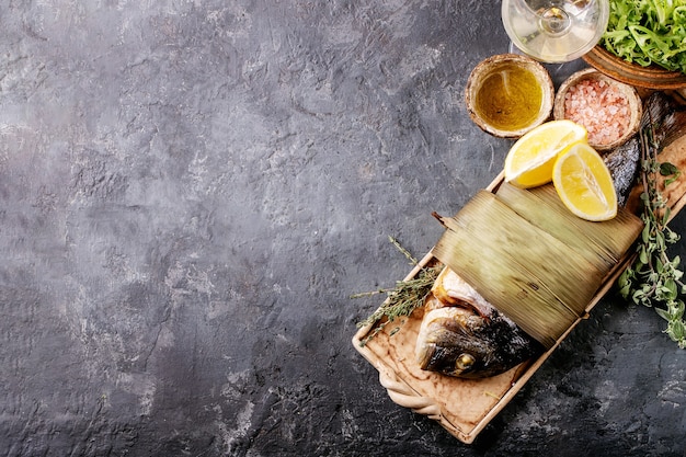 Grilled cooked fresh gutted sea bream or dorado fish served with lemon, green salad, rosemary, olive oil and pink salt on ceramic plate wrapped in bamboo leaves over dark texture surface. Flat lay