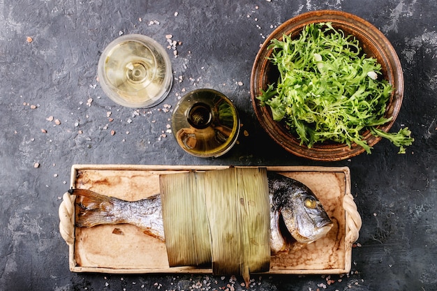 Grilled cooked fresh gutted sea bream or dorado fish served with lemon, green salad, rosemary, olive oil, white wine and pink salt on ceramic plate wrapped in bamboo leaves over dark surface.