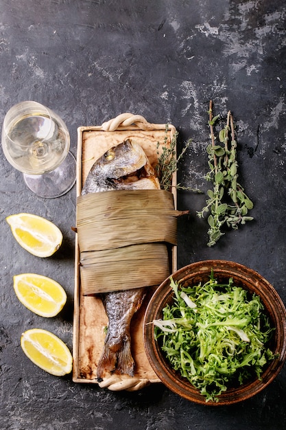 Grilled cooked fresh gutted sea bream or dorado fish served with lemon, green salad, rosemary, olive oil, white wine and pink salt on ceramic plate wrapped in bamboo leaves over dark surface.