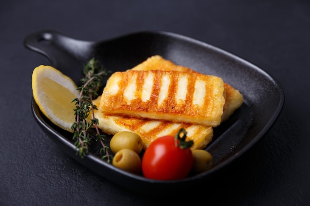 Grilled halloumi cheese in a black pan with olives tomatoes cucumbers thyme and lemon Traditional cheese of the Eastern Mediterranean Black background selective focus closeup