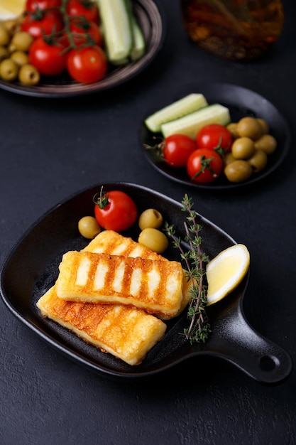 Grilled halloumi cheese in a black pan with olives tomatoes cucumbers thyme and lemon Traditional cheese of the Eastern Mediterranean Black background selective focus closeup