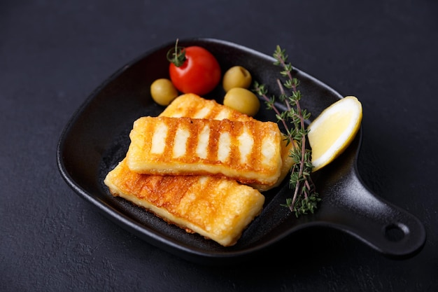 Grilled halloumi cheese in a black pan with olives tomatoes cucumbers thyme and lemon Traditional cheese of the Eastern Mediterranean Black background selective focus closeup