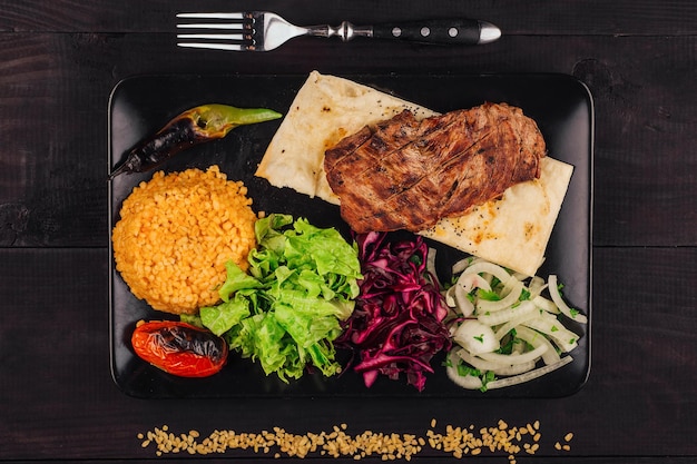 Grilled meat dish served on black plate with vegetables and bulgur