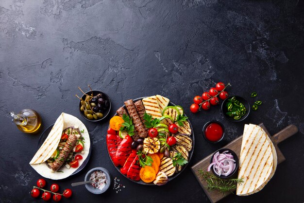 Grilled meat kebabs vegetables on a black plate with tortillas flat bread Slate background Copy space Top view