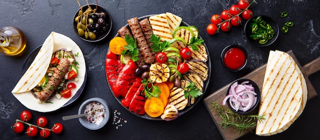 Grilled meat kebabs vegetables on a black plate with tortillas flat bread Slate background Top view